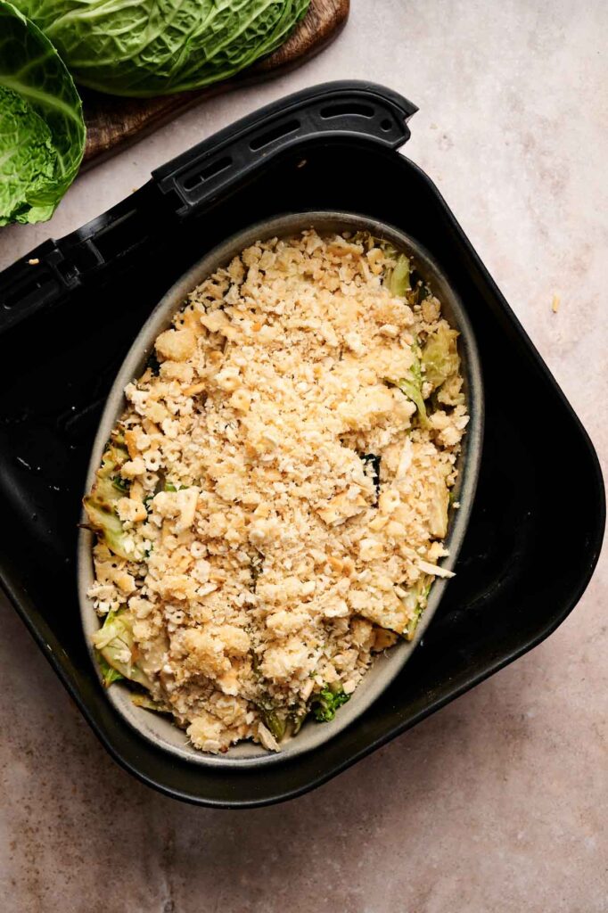 An oval dish of Air Fryer Cabbage Casserole with a golden crumb topping sits in an air fryer basket, while leafy greens peek from the corner on a light countertop.