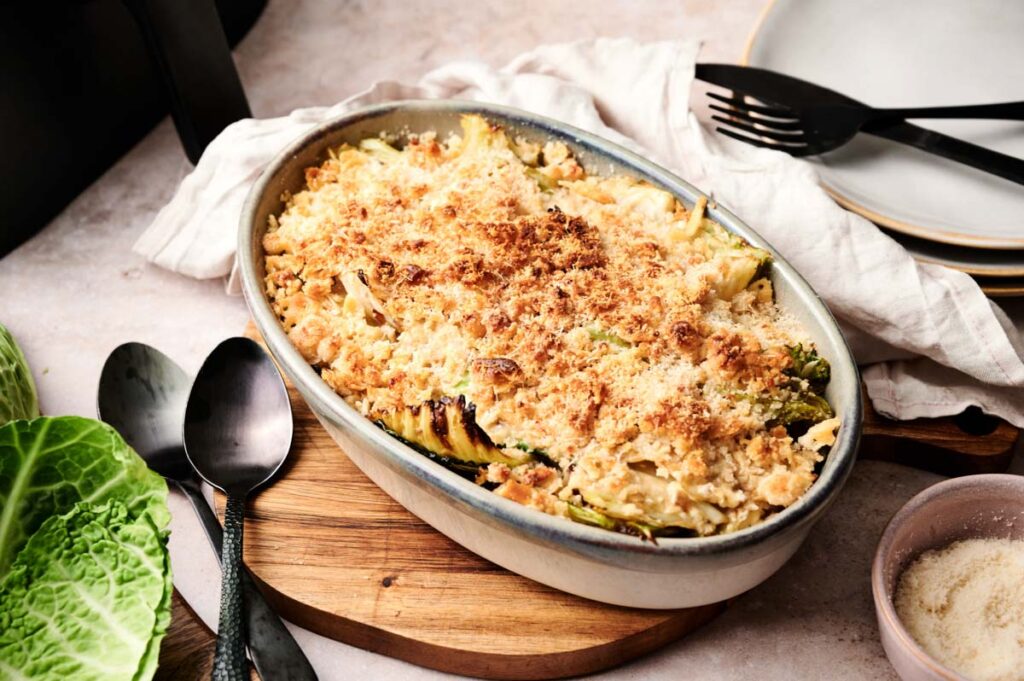 Oval baking dish filled with Air Fryer Cabbage Casserole topped with golden breadcrumbs, set on a wooden board next to serving utensils and a bowl of grated cheese.