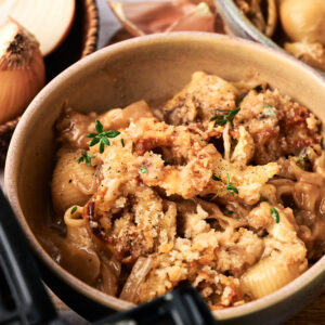 A bowl of pasta shells topped with a golden breadcrumb crust, garnished with fresh herbs, surrounded by onions and a bowl of additional pasta&mdash;perfect for inspiring your next Air Fryer French Onion Casserole.