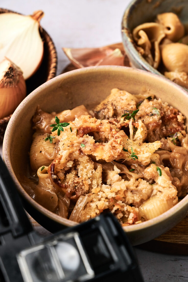 A bowl of pasta shells topped with a golden breadcrumb crust, garnished with fresh herbs, surrounded by onions and a bowl of additional pasta—perfect for inspiring your next Air Fryer French Onion Casserole.