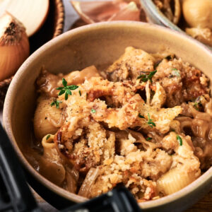 A bowl of pasta shells topped with a golden breadcrumb crust, garnished with fresh herbs, surrounded by onions and a bowl of additional pasta&mdash;perfect for inspiring your next Air Fryer French Onion Casserole.