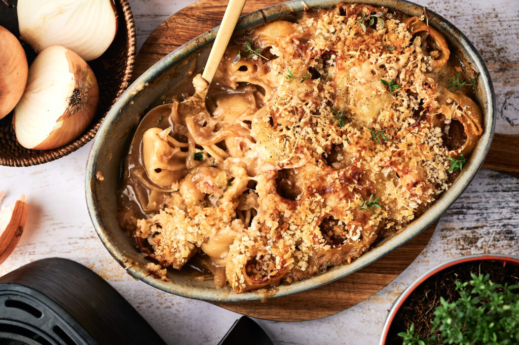 A baked Air Fryer French Onion Casserole filled with savory pasta, topped with golden breadcrumbs and herbs; a wooden spoon rests inside. Sliced onions and a basket of whole onions are nearby.