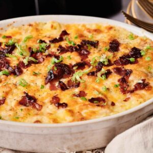 An Air Fryer Twice Baked Potato Casserole topped with melted cheese, crispy bacon pieces, and chopped green onions in a round ceramic dish.