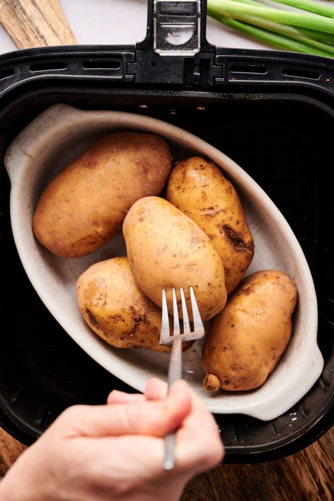 A hand uses a fork to poke holes in raw potatoes placed in a white dish inside an air fryer basket, prepping them for a delicious Air Fryer Twice Baked Potato Casserole.