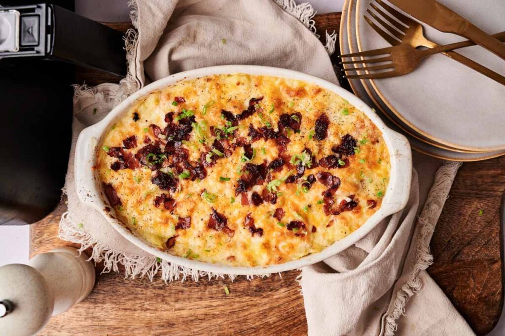 Air Fryer Twice Baked Potato Casserole topped with melted cheese and bacon bits, garnished with herbs, served in a white oval dish on a wooden surface alongside plates and gold utensils.