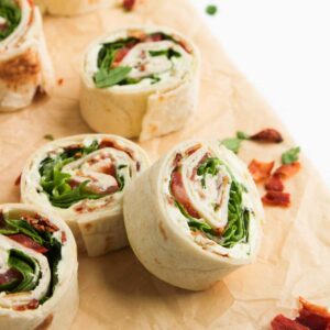 BLT Pinwheels filled with lettuce, tomato, bacon, and a creamy spread are arranged on parchment paper, surrounded by scattered fresh greens.