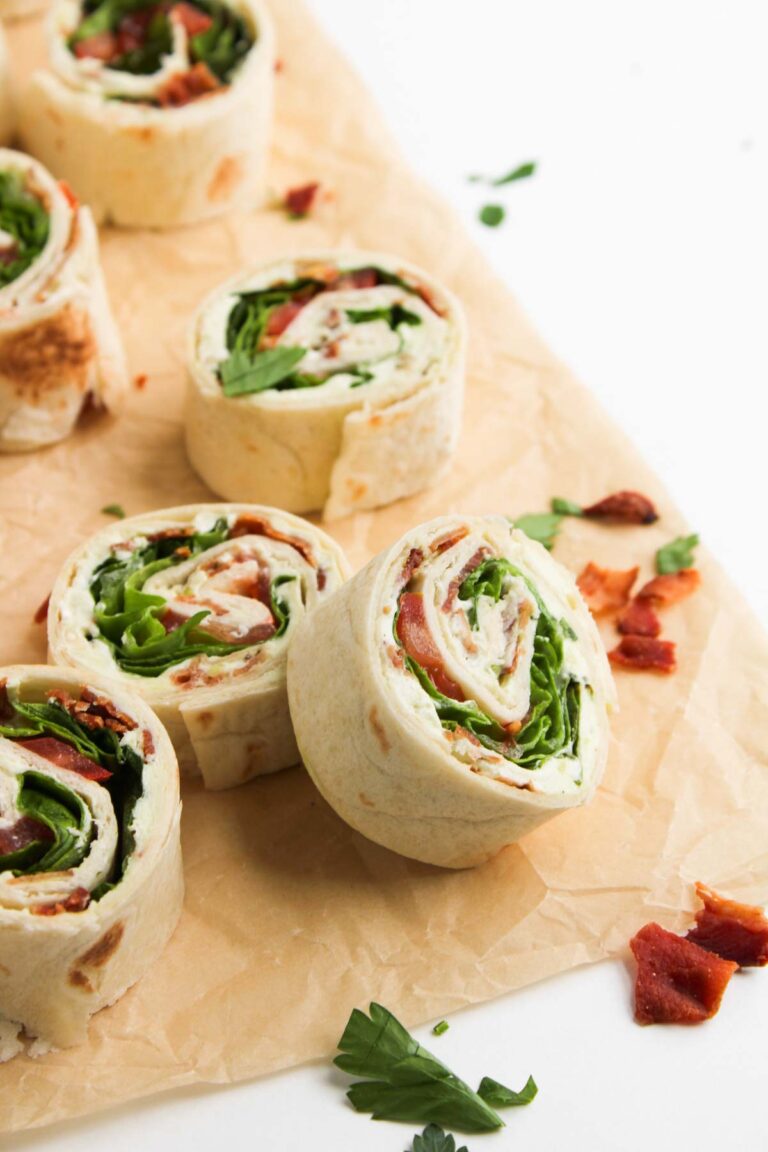 BLT Pinwheels filled with lettuce, tomato, bacon, and a creamy spread are arranged on parchment paper, surrounded by scattered fresh greens.