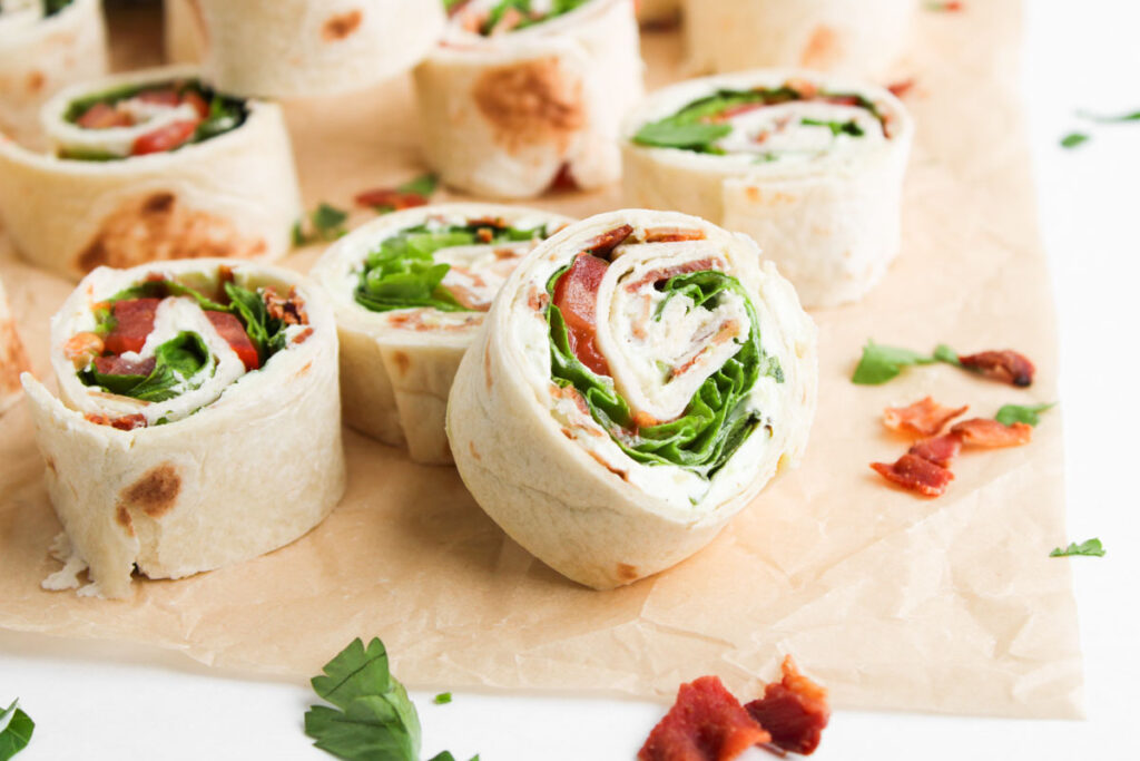 BLT Pinwheels feature sliced tortilla wraps filled with lettuce, bacon, and cream cheese, arranged on parchment paper and garnished with parsley and crispy bacon pieces.