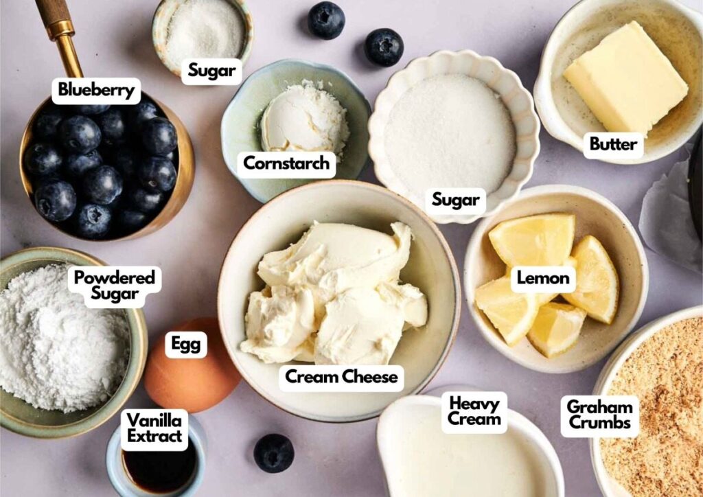 An assortment of ingredients in bowls and plates, perfect for making Air Fryer Blueberry Cheesecake, including blueberries, sugar, cornstarch, butter, lemon, eggs, cream cheese, powdered sugar, vanilla extract, heavy cream, and graham crumbs.