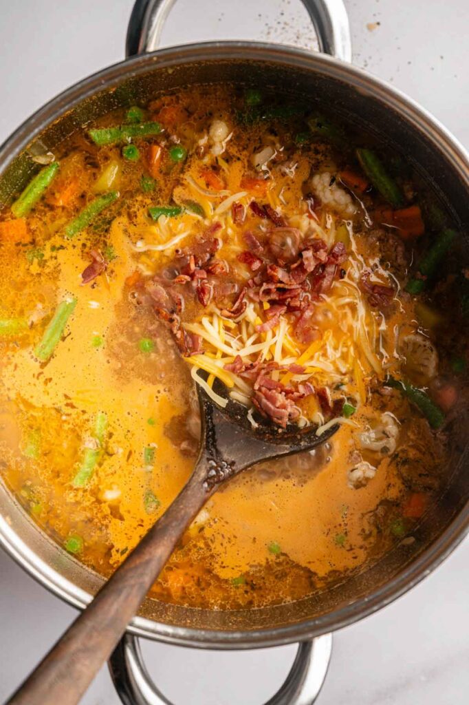 A pot of Cheeseburger Soup with a metal ladle, topped with shredded cheese and bacon bits, contains vegetables and a creamy broth.