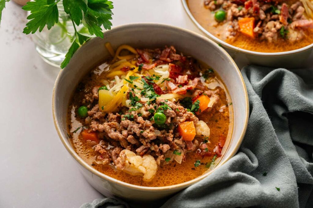 A bowl of Cheeseburger Soup with ground meat, peas, carrots, cauliflower, shredded cheese, and chopped herbs, with another bowl and a parsley garnish in the background.