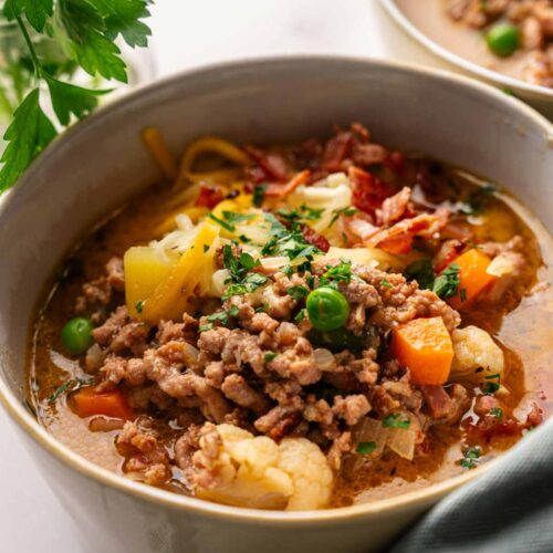 A hearty bowl of Cheeseburger Soup with ground meat, peas, carrots, potatoes, cauliflower, and herbs, garnished with chopped parsley.
