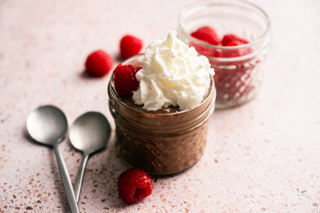 A small glass jar of Cottage Cheese Chocolate Mousse topped with whipped cream and raspberries, with another jar of raspberries and two metal spoons beside it.