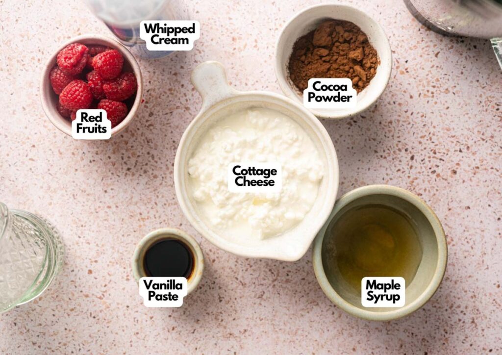 A flat lay of ingredients for Cottage Cheese Chocolate Mousse, including whipped cream, cocoa powder, cottage cheese, red fruits, vanilla paste, and maple syrup, all arranged on a speckled countertop.