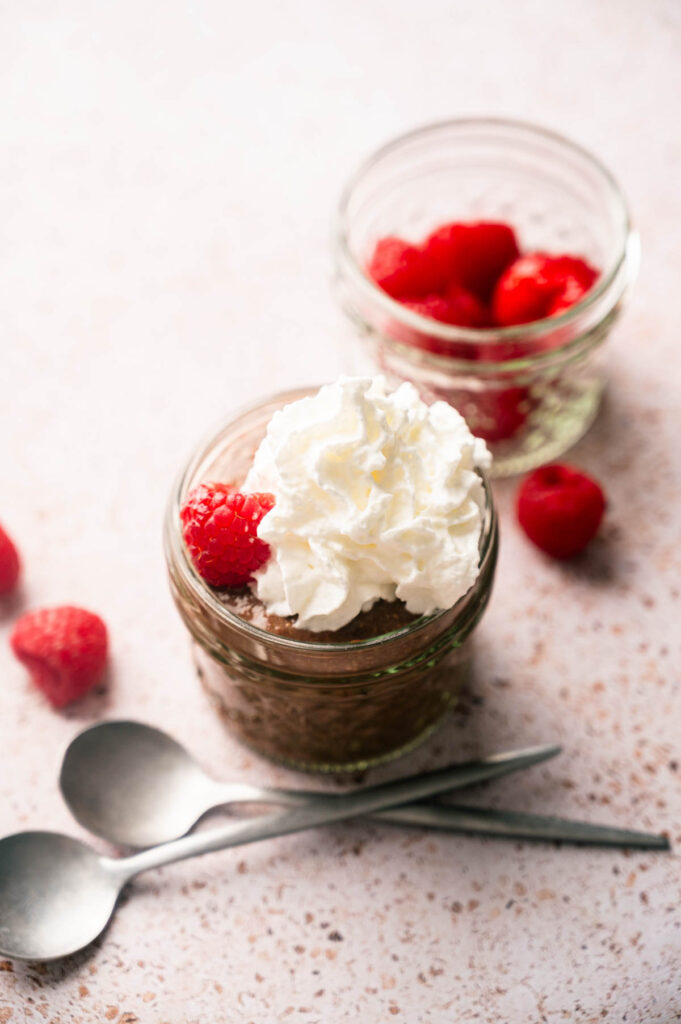 A small glass jar filled with Cottage Cheese Chocolate Mousse, topped with whipped cream and a raspberry, sits beside another jar containing raspberries, with two spoons and loose raspberries nearby.