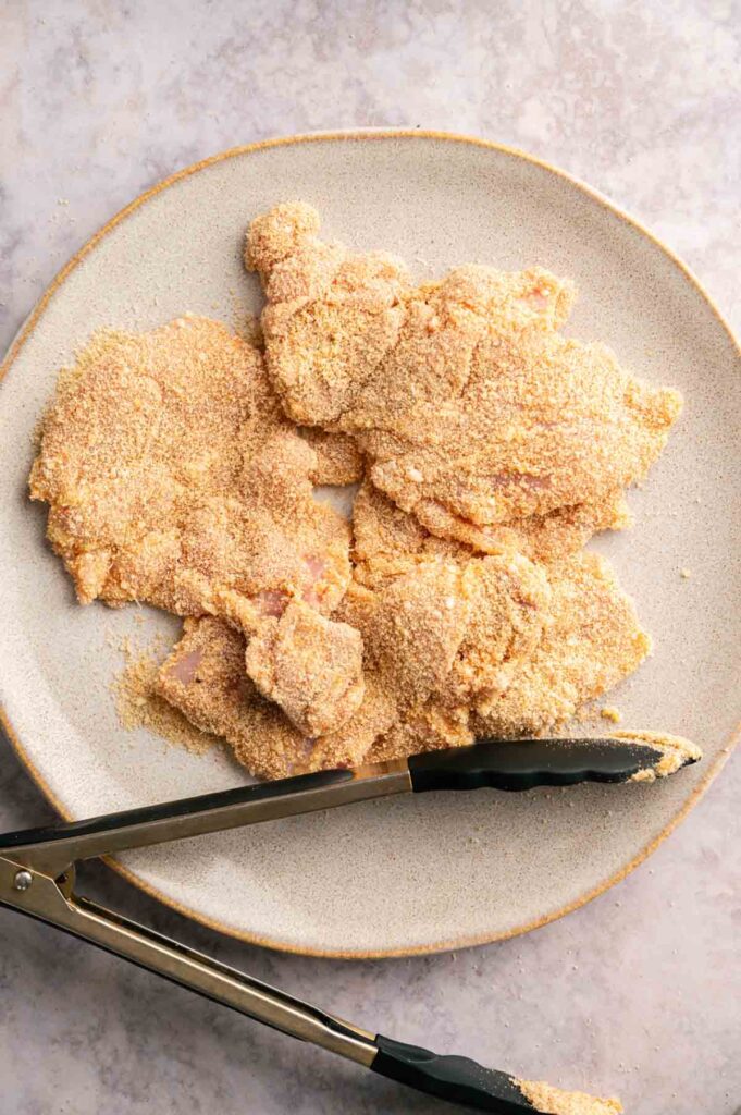 Plate with raw chicken pieces coated in breadcrumbs for Crispy Chicken Thighs, with metal tongs resting on the edge of the plate, on a light-colored surface.