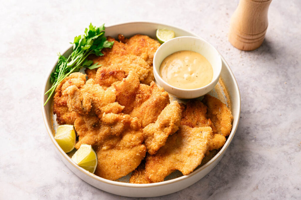 A plate of crispy chicken thighs breaded and fried, served with lime wedges, fresh parsley, and a bowl of dipping sauce.