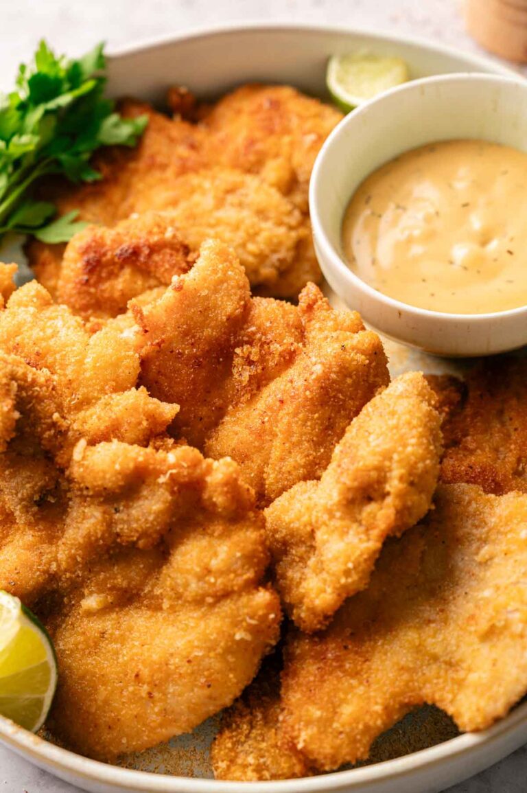 Crispy chicken thighs breaded and fried to perfection, served with a bowl of creamy dipping sauce, lime wedges, and fresh parsley.