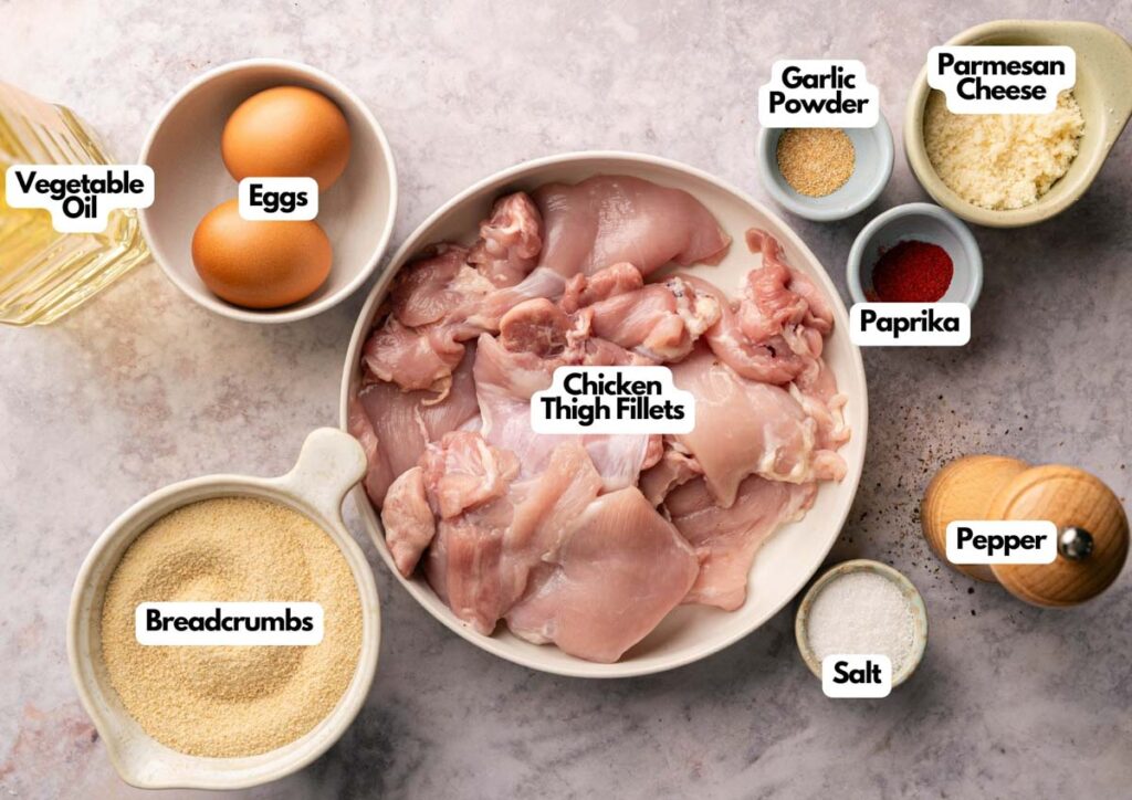 A bowl of raw chicken thigh fillets sits surrounded by breadcrumbs, eggs, garlic powder, parmesan, and spices—everything you need to create deliciously crispy chicken thighs right on your countertop.