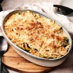 An oval baking dish filled with golden-brown, breadcrumb-topped Air Fryer Cabbage Casserole sits on a wooden board next to a spoon and plates.