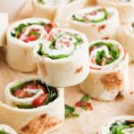 Several BLT Pinwheels filled with lettuce, tomato, and a creamy spread are arranged on parchment paper.