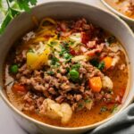 A bowl of Cheeseburger Soup with ground meat, cauliflower, peas, carrots, shredded cheese, bacon bits, and chopped herbs is served on a table.