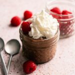 A small glass jar filled with Cottage Cheese Chocolate Mousse, topped with whipped cream and raspberries; another jar with raspberries and two spoons are beside it.