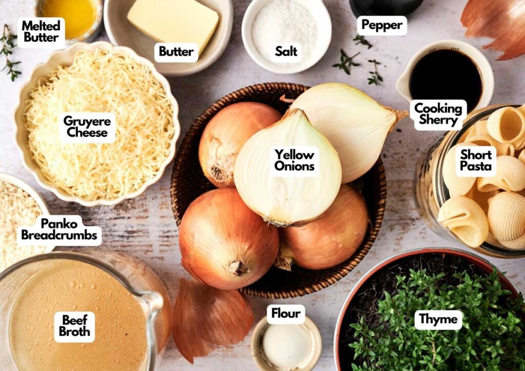 A variety of labeled ingredients for an Air Fryer French Onion Casserole are arranged on a table, including yellow onions, cheese, butter, pasta, broth, flour, breadcrumbs, sherry, thyme, salt, and pepper.