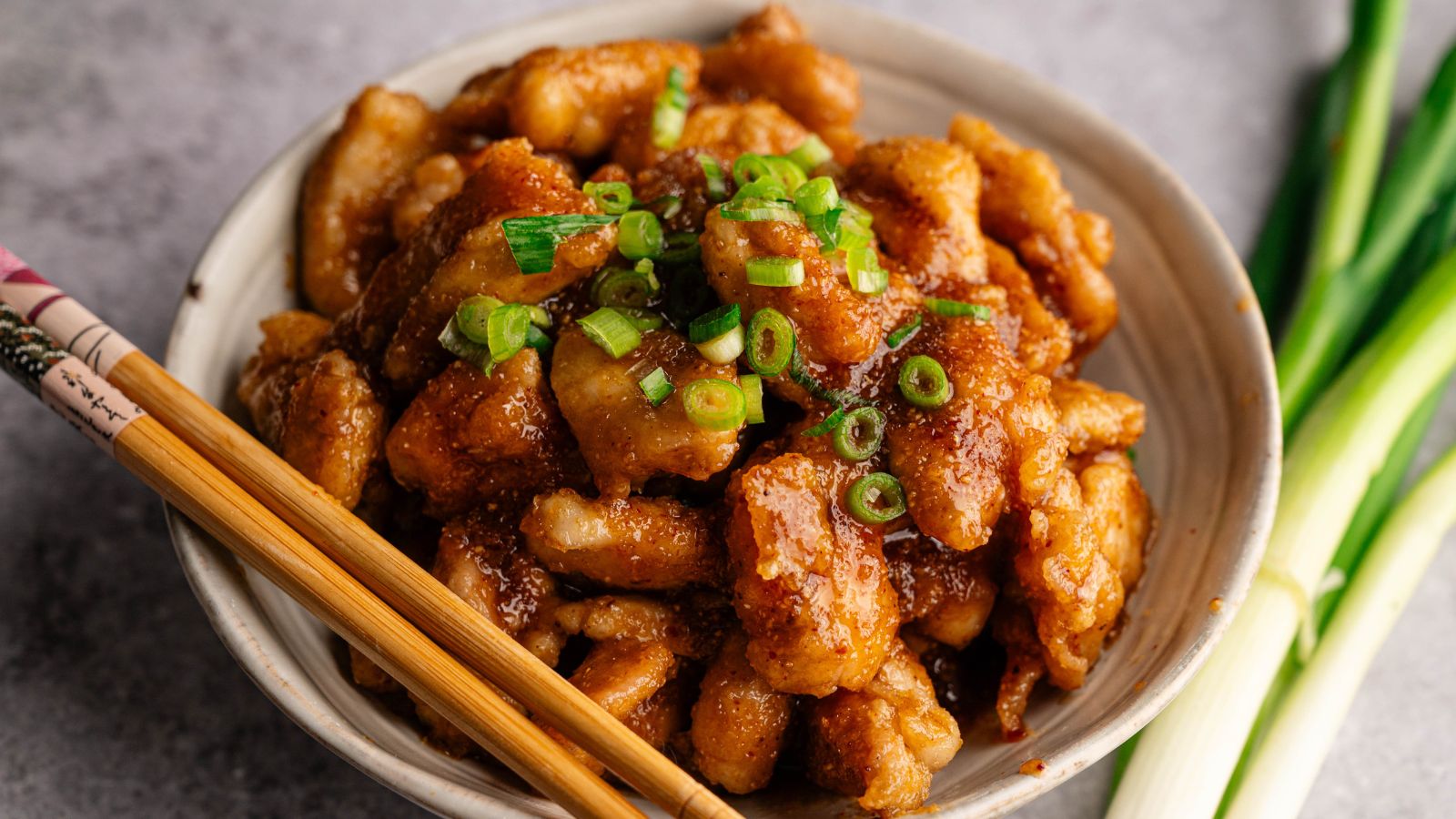 A bowl of glazed chicken pieces topped with chopped green onions, with chopsticks resting on the bowl and green onions beside it.