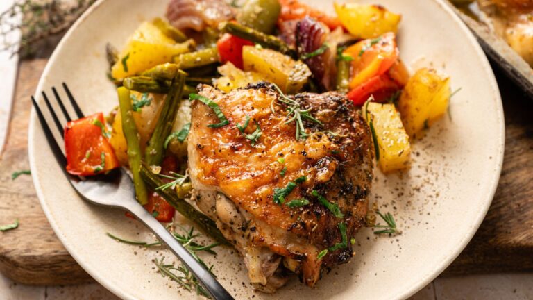 A plate with roasted chicken thigh, garnished with herbs, served alongside roasted vegetables including potatoes, green beans, red onions, and bell peppers. A fork rests on the plate.