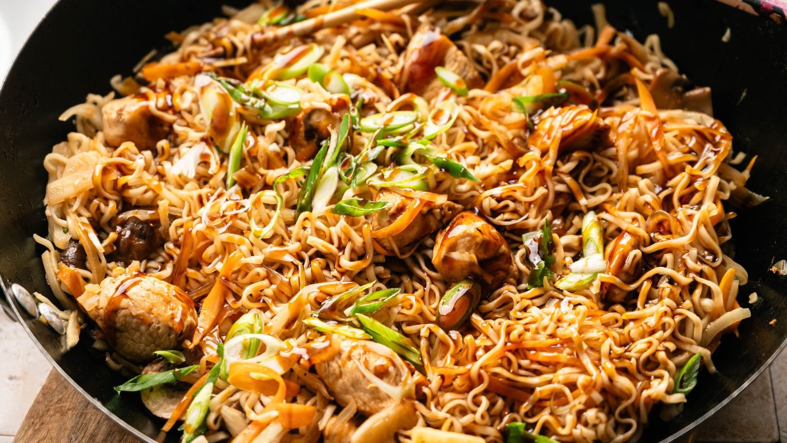 A close-up of cooked stir-fried noodles with pieces of chicken, sliced carrots, green onions, and a savory sauce in a black pan.