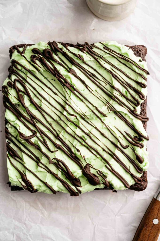 A square Mint Chocolate Brownie topped with light green mint frosting and drizzled with dark chocolate, placed on white parchment paper.