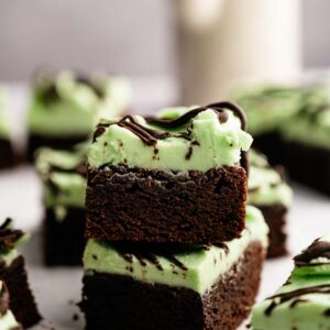 Two Mint Chocolate Brownies with a layer of green mint frosting and a drizzle of chocolate on top, stacked on parchment paper.