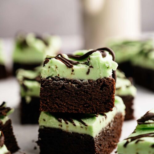 Two Mint Chocolate Brownies with a layer of green mint frosting and a drizzle of chocolate on top, stacked on parchment paper.
