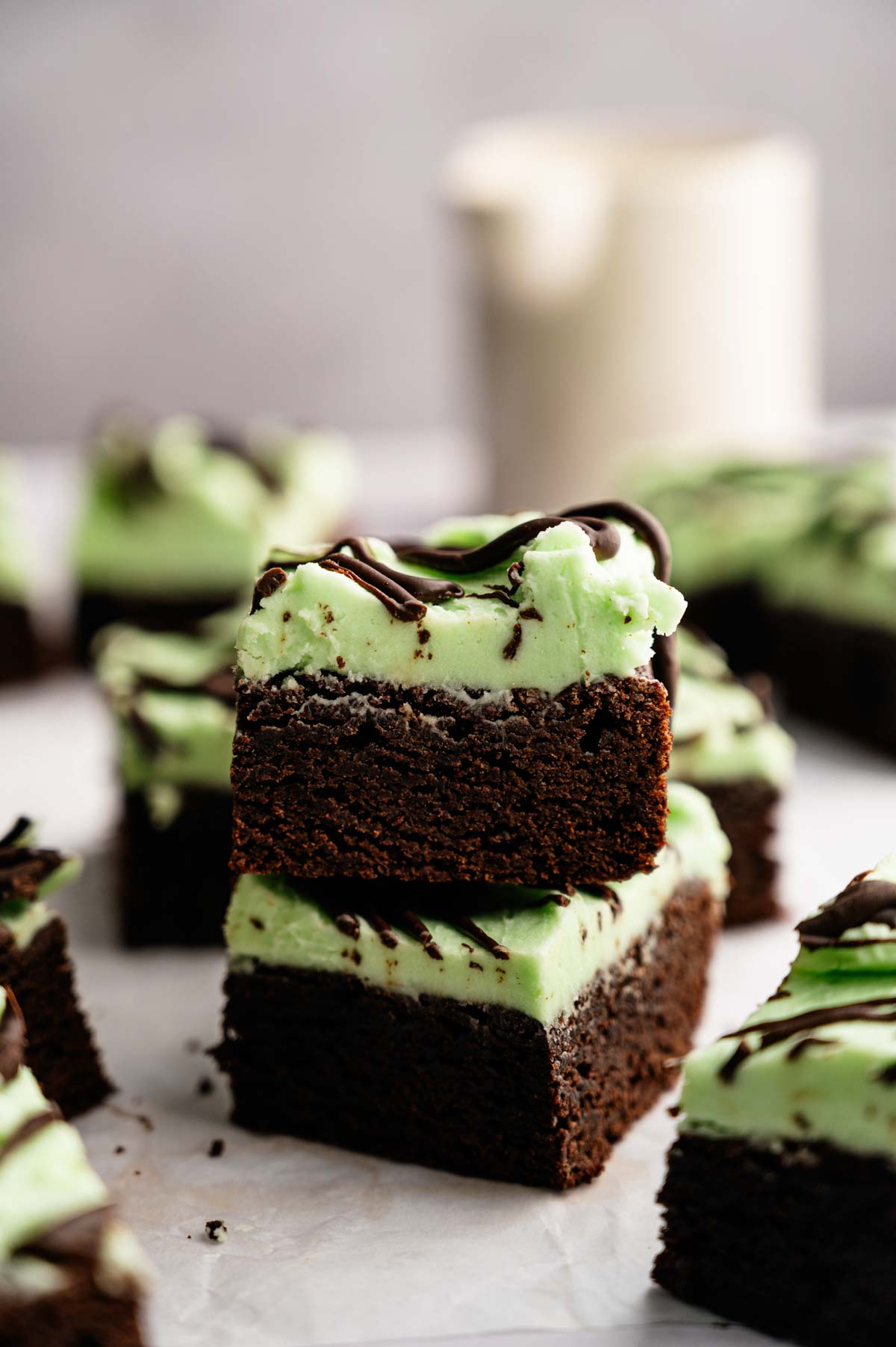 Two Mint Chocolate Brownies with a layer of green mint frosting and a drizzle of chocolate on top, stacked on parchment paper.