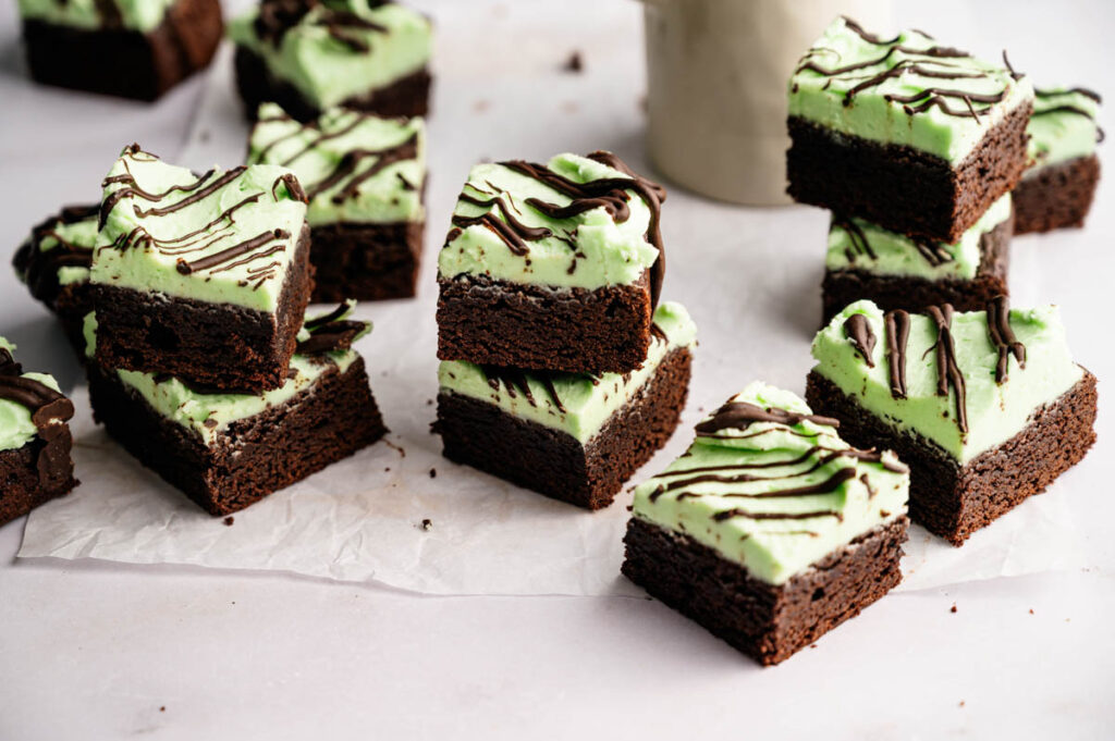 Squares of Mint Chocolate Brownies topped with green mint frosting and a chocolate drizzle, arranged in small stacks on parchment paper.