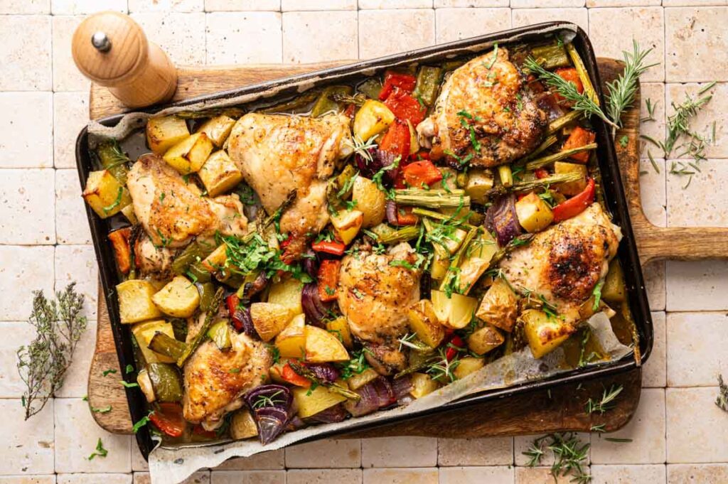 A baking tray of Sheet Pan Chicken Thighs and Veggies&mdash;roasted chicken thighs, potatoes, peppers, and onions with fresh herbs&mdash;rests on a tiled surface beside a wooden pepper grinder.