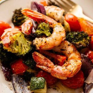 A plate of Sheet Pan Shrimp with roasted broccoli, zucchini, tomatoes, and red onion, seasoned with herbs and served with a fork.