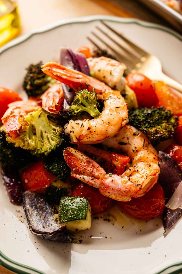 A plate of Sheet Pan Shrimp with roasted broccoli, zucchini, tomatoes, and red onion, seasoned with herbs and served with a fork.