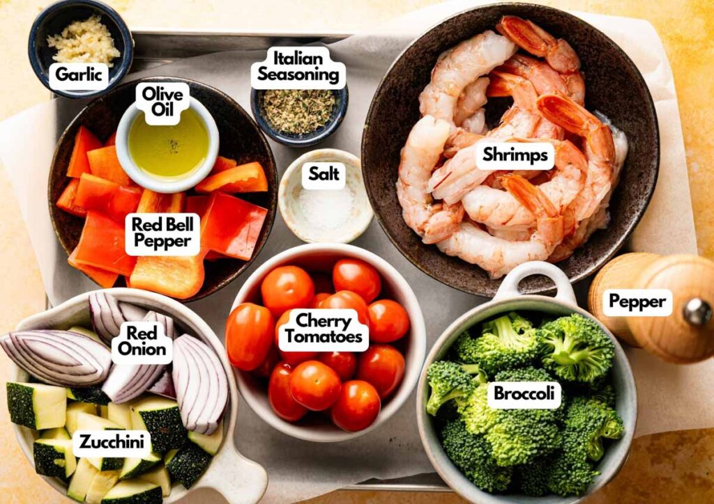 A tray with bowls containing ingredients for Sheet Pan Shrimp: shrimp, broccoli, cherry tomatoes, red onion, zucchini, red bell pepper, garlic, olive oil, salt, pepper, and Italian seasoning.