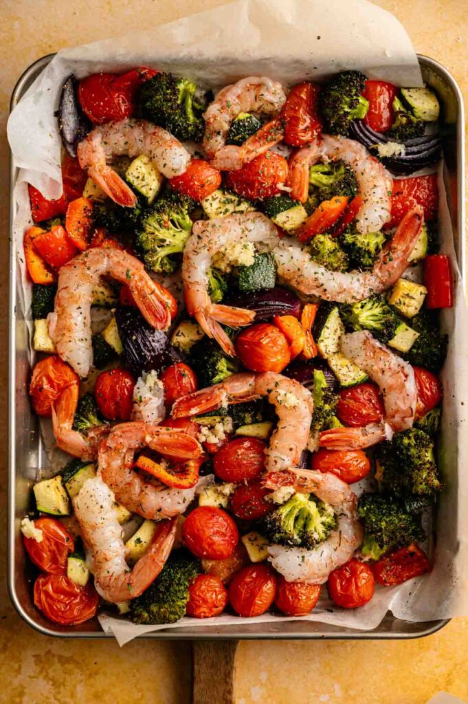 A sheet pan shrimp dinner with raw shrimp, cherry tomatoes, broccoli, red onion, zucchini, and bell peppers seasoned with herbs&mdash;all ready to be roasted for a quick and colorful meal.