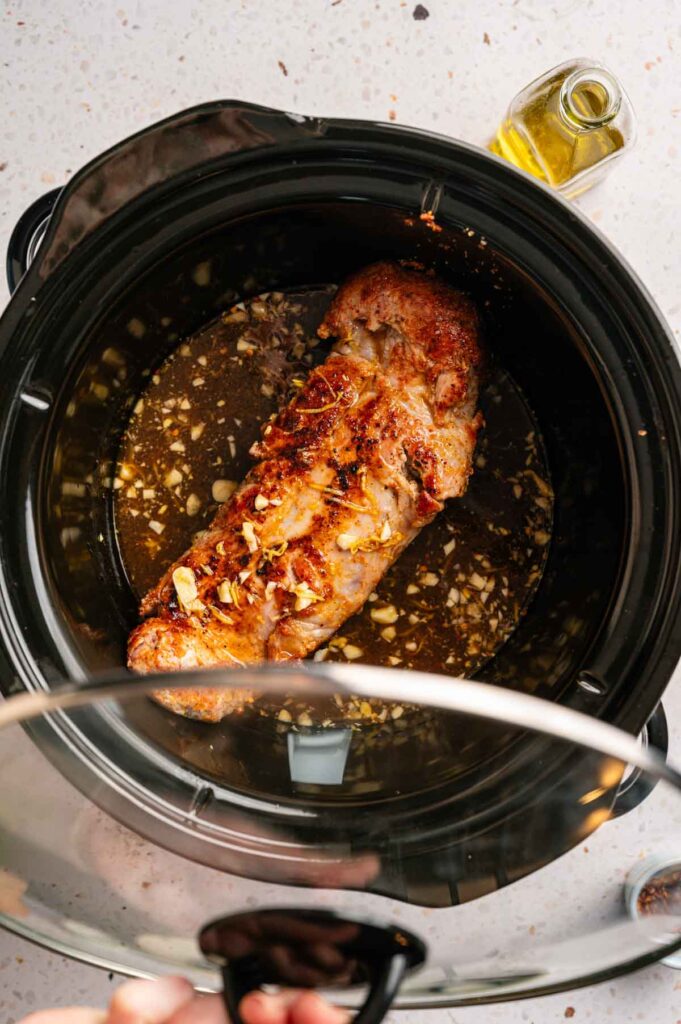 A seasoned Slow Cooker Pork Tenderloin rests in its juices as someone lifts the glass lid, revealing the tender meat. A small bottle of oil sits nearby on the countertop.