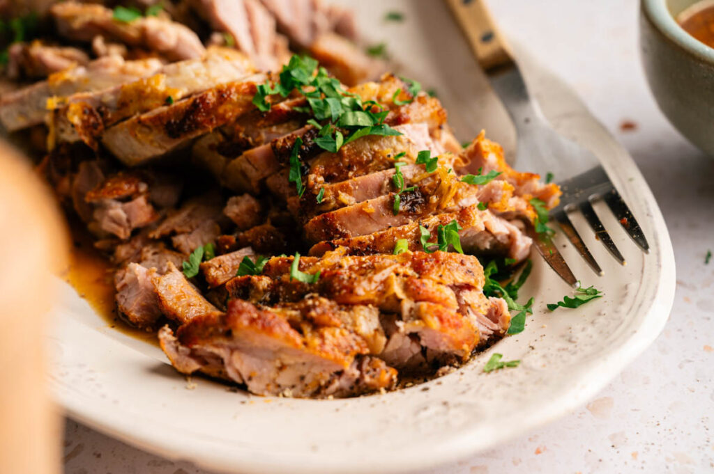 Slow Cooker Pork Tenderloin sliced and garnished with chopped herbs on a white platter, with a fork beside the meat.
