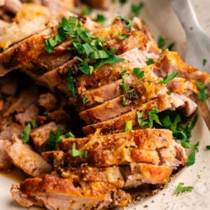 Slow Cooker Pork Tenderloin, sliced and roasted to perfection, is garnished with chopped fresh herbs on a white serving platter, with a serving knife nearby.