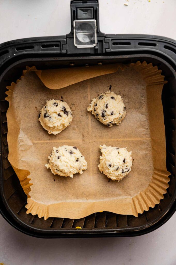 Four scoops of Air Fryer Cannoli Cookies dough are placed on parchment paper inside an air fryer basket, ready to be baked.