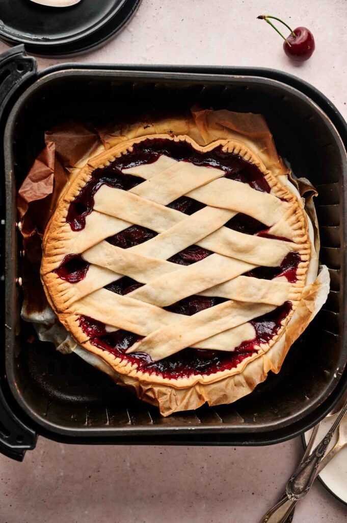 An Air Fryer Cherry Pie with a golden lattice crust sits on parchment paper inside an air fryer basket, with a single cherry and utensils nearby.