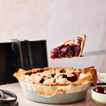 A slice of Air Fryer Cherry Pie is being lifted from a pie dish, revealing a thick cherry filling and lattice crust; a fresh cherry and pie server are nearby.