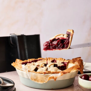 A slice of Air Fryer Cherry Pie is being lifted from a pie dish, revealing a thick cherry filling and lattice crust; a fresh cherry and pie server are nearby.