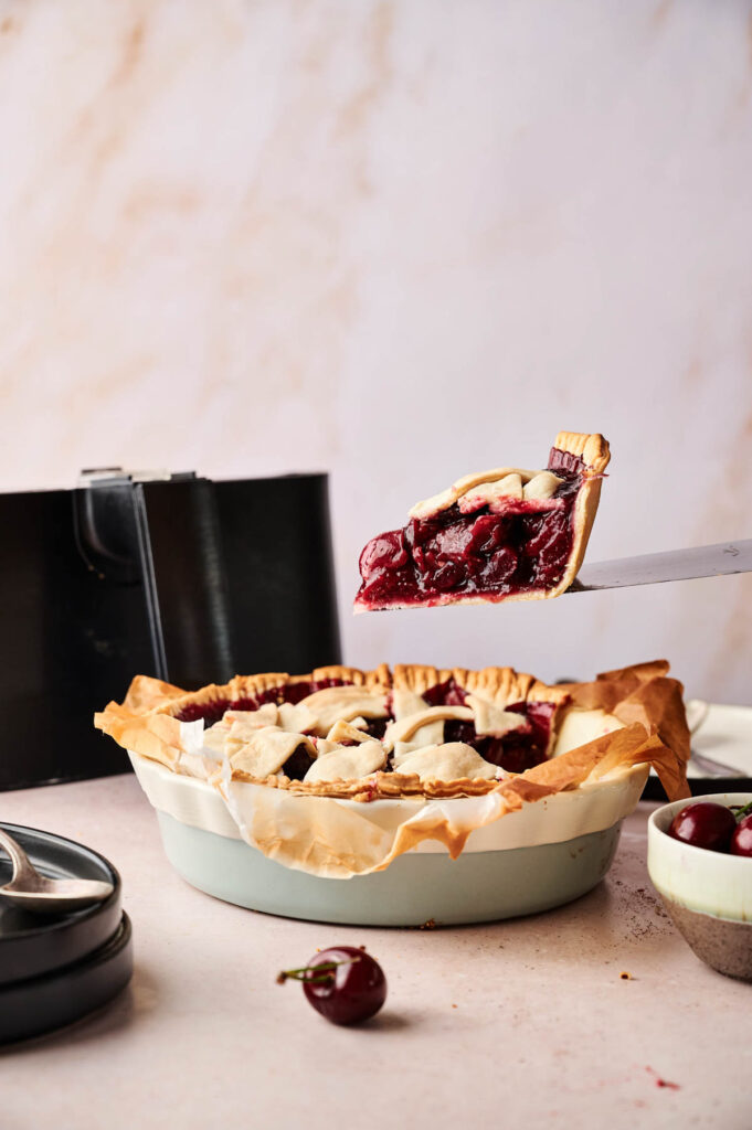 A slice of Air Fryer Cherry Pie is being lifted from a pie dish, revealing a thick cherry filling and lattice crust; a fresh cherry and pie server are nearby.