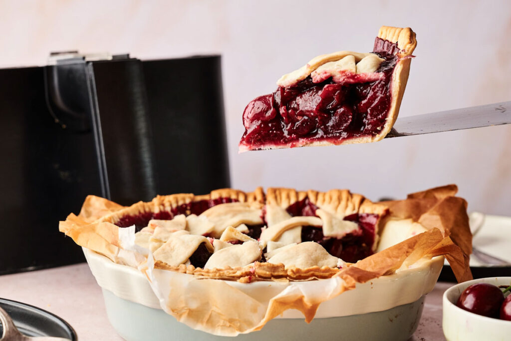 A slice of Air Fryer Cherry Pie is being lifted from a pie dish lined with parchment paper, revealing a luscious filling of whole cherries and a perfectly golden lattice crust.